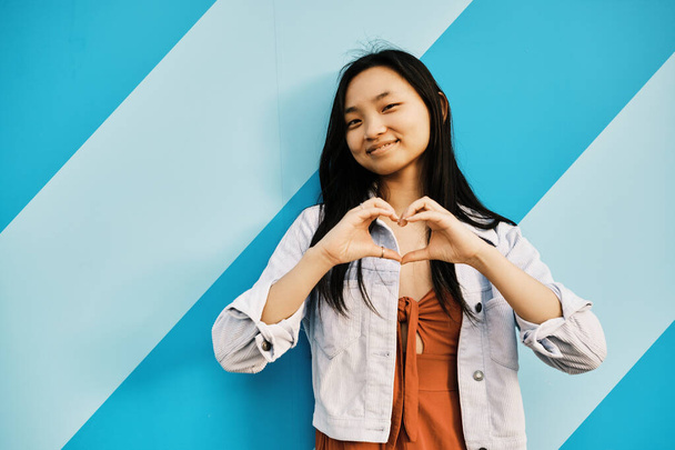 Uśmiechnięta młoda Azjatka kształtująca serce dłońmi stojąc przy ścianie w paski. Pojęcie miłości. - Zdjęcie, obraz