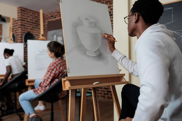 Sanat dersi sırasında resim çiziminde çalışan yaratıcı öğrenci beyaz tuvalin önünde vazo modeli çiziyor. Çeşitli takımlar yaratıcılık stüdyosunda skeç dersine katılıyor. - Fotoğraf, Görsel