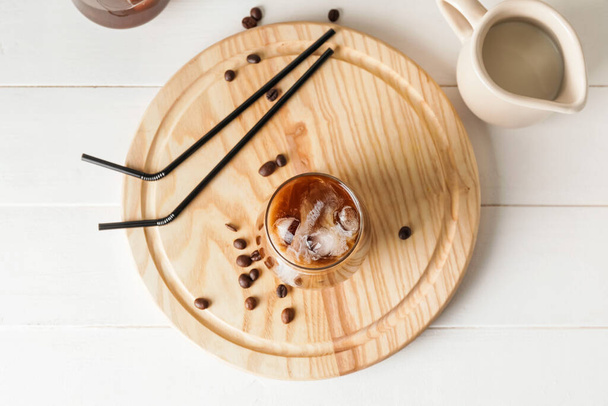 Drewniana deska ze szklanką zimnej kawy, mleka i słomek na białym stole - Zdjęcie, obraz