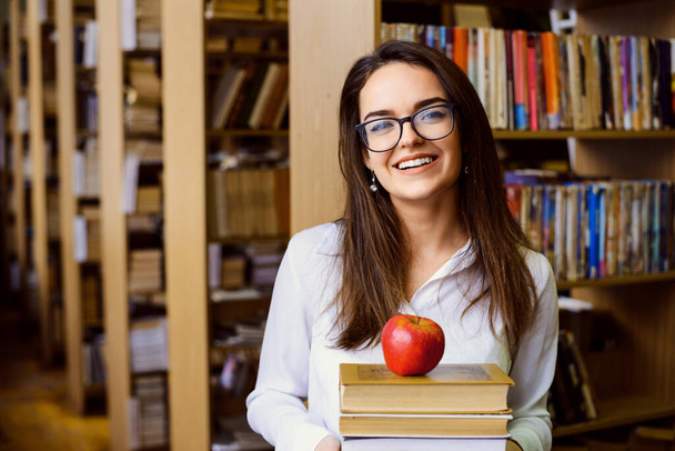 Szczęśliwy student z mnóstwem książek i jabłkiem w bibliotece. Ciężko pracujący student lubi się uczyć. - Zdjęcie, obraz