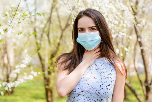 Mujer joven en máscara médica que tiene dolor de garganta y alergia de pie al aire libre contra los árboles en flor - Foto, Imagen