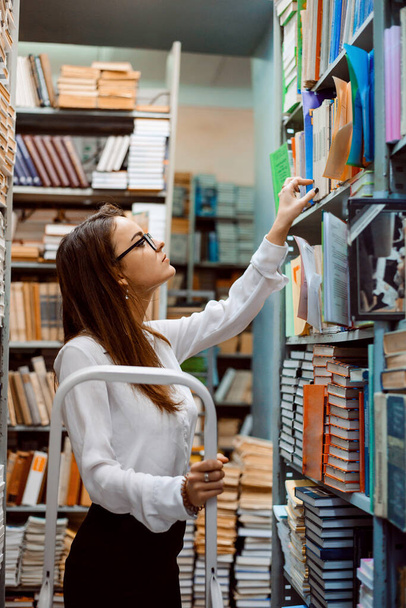 Junge attraktive Bibliothekarin steht auf der Leiter und überprüft Bücher, ihren Zustand und ihre Präsenz - Foto, Bild
