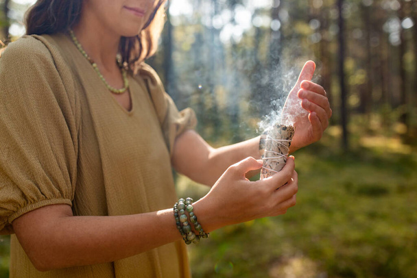occult science and supernatural concept - close up of woman or witch with smoking white sage performing magic ritual in forest - Foto, imagen