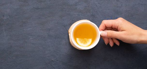 Female hand holding a cup of tea with lemon smile top view on a dark stone background. - 写真・画像
