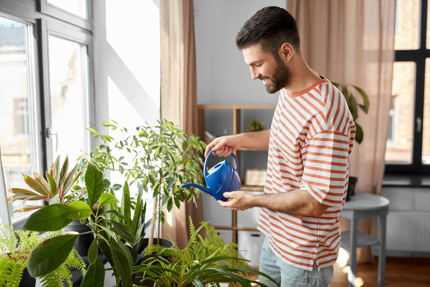 people, nature and plants care concept - happy smiling man watering flowers at home - Foto, imagen