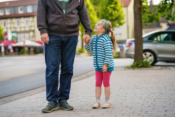 Fröhliches Vorschulmädchen lächelt und hält Väter auf dem Schulweg am ersten sonnigen Schultag an der Hand. Vater und Tochter auf dem Weg in den Kindergarten, Spaß zusammen. Kind mit Brille - Foto, Bild
