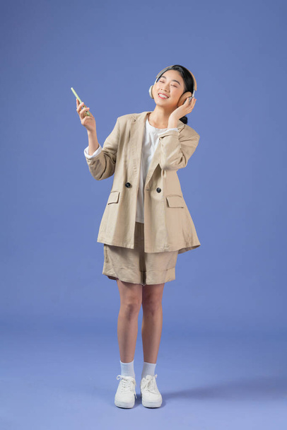 Χαρούμενη όμορφη νεαρή Ασιάτισσα γυναίκα ακούγοντας μουσική με ασύρματα ακουστικά που απομονώνονται σε μωβ φόντο, κρατώντας το κινητό τηλέφωνο - Φωτογραφία, εικόνα