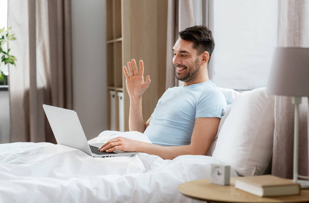 technology, people and lifestyle concept - happy man with laptop computer having video call in bed at home bedroom - Foto, imagen