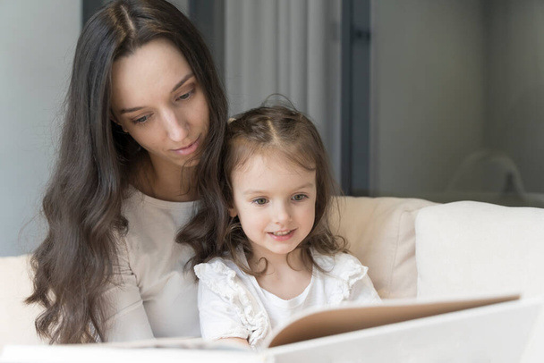 Mom and daughter are reading a book on the sofa. Study and activities with children at home, mental development, free time with children. High quality photo - Φωτογραφία, εικόνα