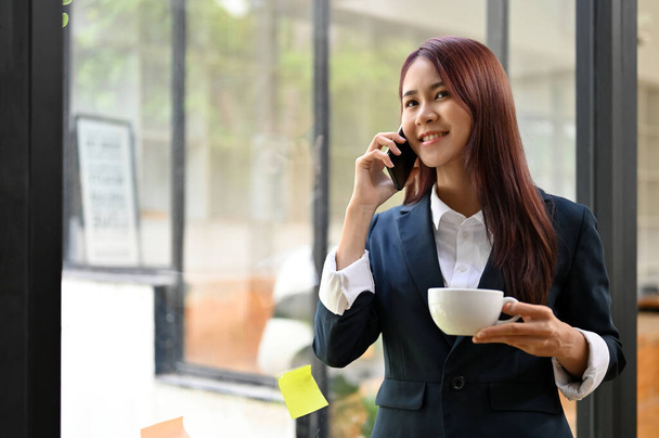 Úspěšná asijská podnikatelka nebo podnikatelka výkonný manažer podnikatel stojící u okna, mluvit do telefonu a popíjet kávu. - Fotografie, Obrázek