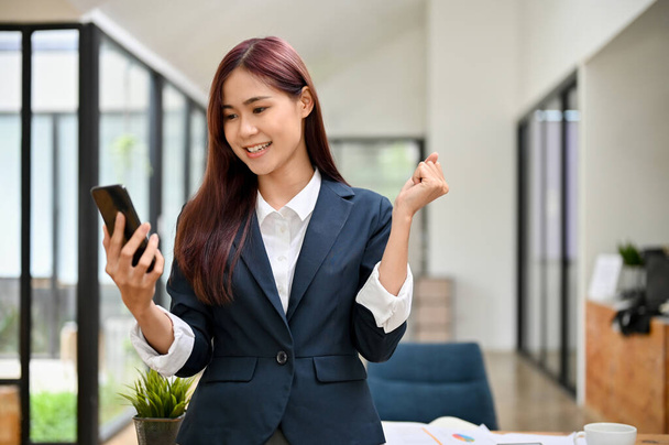 Alegre sorprendido exitoso joven mujer de negocios asiática de pie en la oficina, utilizando un teléfono inteligente, recibiendo una noticia sorprendida. - Foto, Imagen