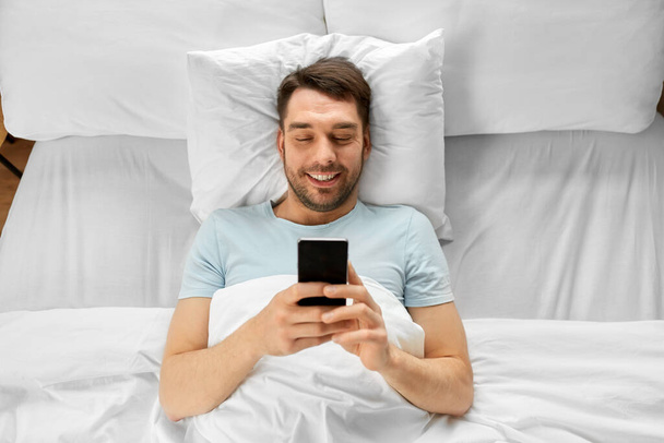 teknologia, internet ja ihmiset käsite hymyilevä mies kutoma älypuhelimeen sängyssä kotona aamulla - Valokuva, kuva