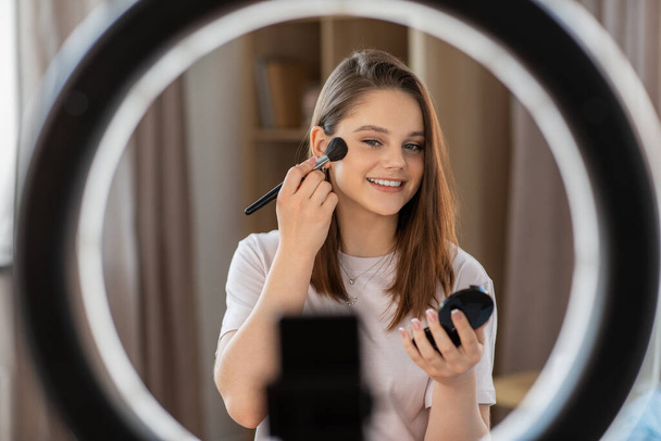 beauty blogging, technology and people concept - happy smiling girl blogger with ring light and smartphone applying make up at home - Foto, afbeelding