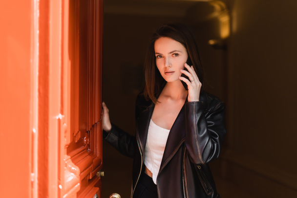 オレンジ色のドアの近くのスマートフォンで話している黒い革のジャケットの若い女性 - 写真・画像