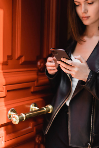 mujer joven en la chaqueta de cuero negro de mensajería en el teléfono inteligente cerca de la puerta naranja - Foto, imagen