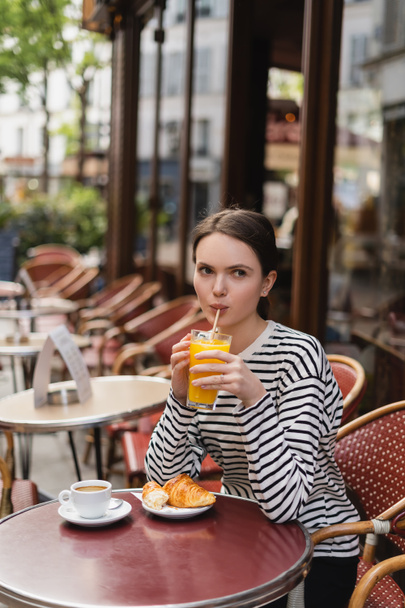 νεαρή γυναίκα πίνει χυμό πορτοκαλιού κοντά στο κρουασάν και καφέ στο τραπέζι στην καλοκαιρινή βεράντα στο γαλλικό καφέ - Φωτογραφία, εικόνα