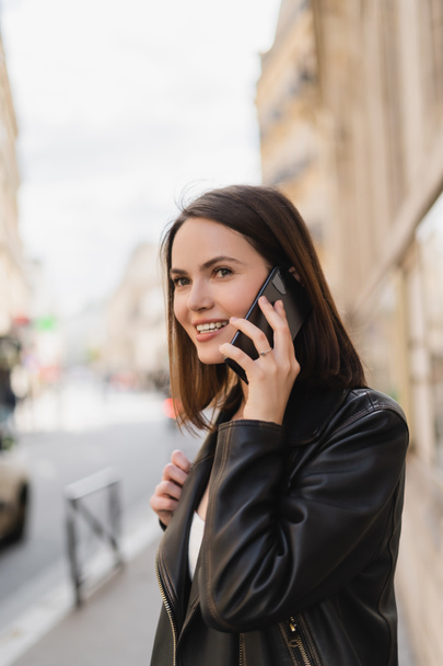 jeune femme positive en veste élégante parlant sur smartphone dans la rue à Paris  - Photo, image