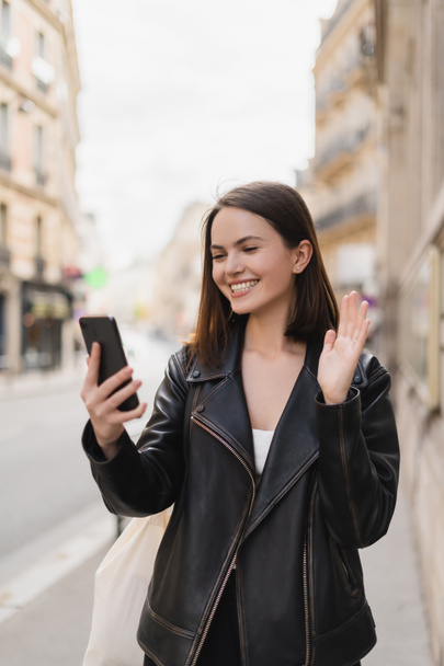 cheerful young woman in stylish jacket waving hand during video chat on street in paris  - Photo, Image