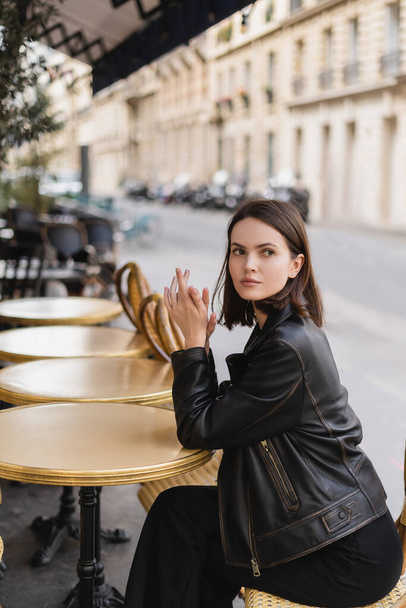 κομψή νεαρή γυναίκα με μαύρο δερμάτινο μπουφάν κάθεται στο γαλλικό καφέ στην καλοκαιρινή βεράντα  - Φωτογραφία, εικόνα