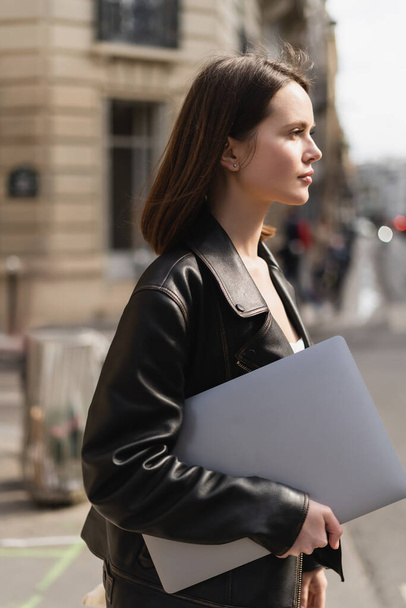 oldalnézetben fiatal szabadúszó fekete bőrdzsekiben séta laptop az utcán Párizsban - Fotó, kép