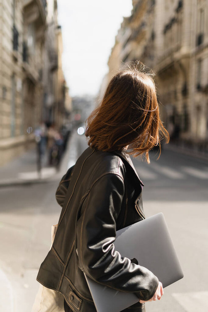 back view of freelancer in black leather jacket walking with laptop on street in paris - Photo, Image