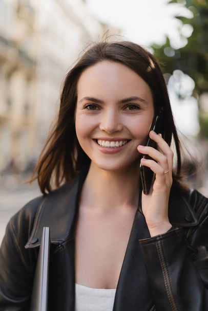 portrait of cheerful woman in black leather jacket holding laptop and talking on smartphone on street in paris - Photo, Image
