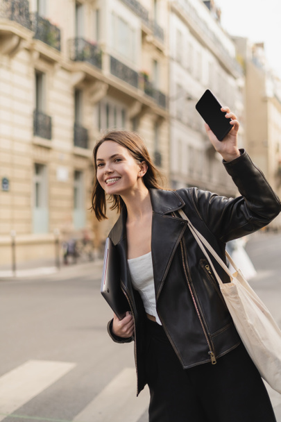 happy woman in black leather jacket holding laptop and waving with smartphone on street in paris - Фото, изображение