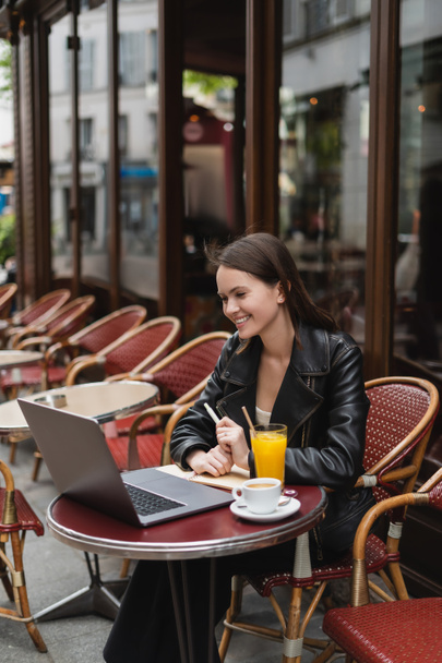 happy freelancer in black leather jacket looking at laptop near drinks on table in french outdoor cafe  - Foto, afbeelding
