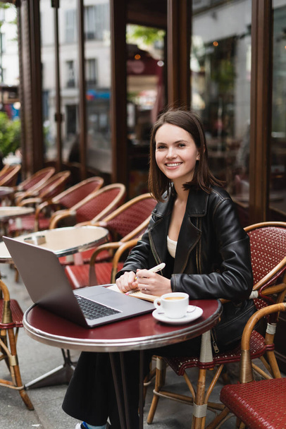 happy freelancer in black jacket looking at camera near laptop and cup of coffee on table in french outdoor cafe - 写真・画像