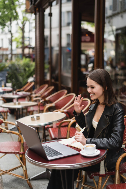 heureux pigiste en veste noire ayant appel vidéo près de tasse de café sur la table dans un café en plein air français  - Photo, image