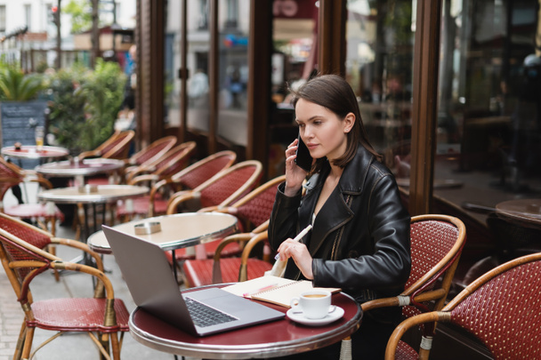 młoda kobieta w czarnej kurtce rozmawia na smartfonie w pobliżu laptopa i filiżanki kawy na stole we francuskiej kawiarni na świeżym powietrzu  - Zdjęcie, obraz