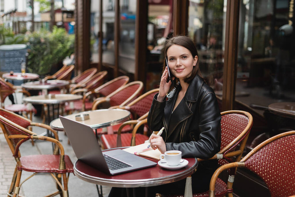 mosolygós szabadúszó fekete kabát beszél okostelefon közelében laptop és csésze kávé az asztalon francia szabadtéri kávézó - Fotó, kép