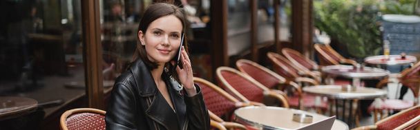 mujer sonriente en chaqueta negra hablando en el teléfono inteligente en la cafetería al aire libre francesa, pancarta - Foto, imagen
