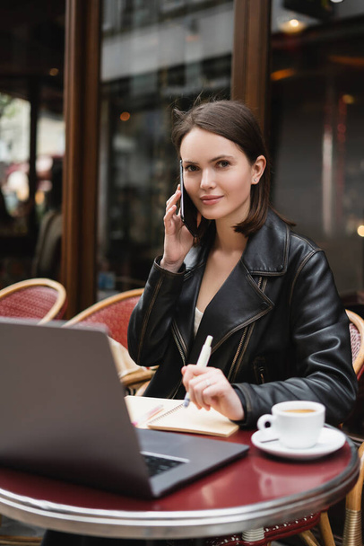 freelancer w czarnej kurtce rozmowy na smartfonie w pobliżu laptopa i filiżanki kawy na stole we francuskiej kawiarni na świeżym powietrzu - Zdjęcie, obraz