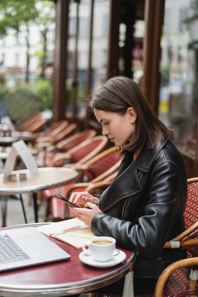 side view of freelancer in black jacket using smartphone near laptop and cup of coffee in french outdoor cafe  - Valokuva, kuva