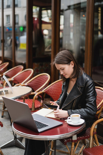 Siyah ceketli genç serbest çalışan bilgisayarın yanında boş ekranlı akıllı telefon ve açık hava kafede bir fincan kahve kullanıyor.  - Fotoğraf, Görsel