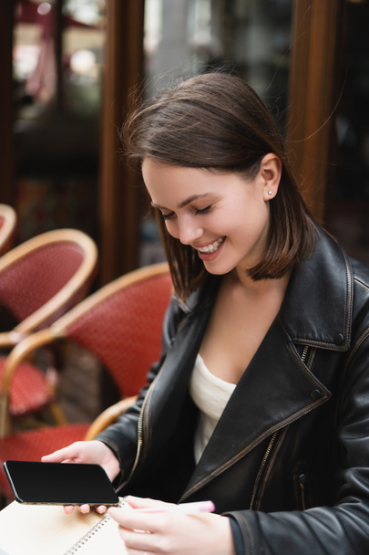 retrato de mujer sonriente sosteniendo teléfono inteligente con pantalla en blanco en la cafetería al aire libre - Foto, imagen