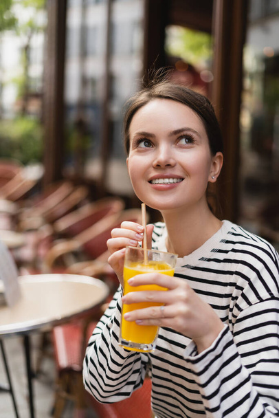 donna allegra in camicia a maniche lunghe a righe con un bicchiere di succo d'arancia fresco in un caffè all'aperto a Parigi - Foto, immagini