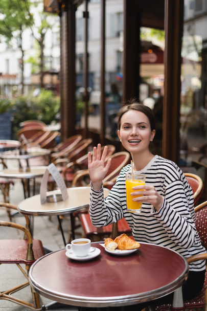 femme heureuse en chemise à manches longues rayée tenant un verre de jus d'orange et agitant la main dans un café en plein air à Paris - Photo, image