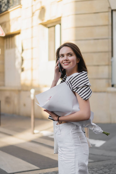 onnellinen nuori nainen tyylikkäässä asussa puhuu älypuhelimella ja pitää kukkakimpun käärittynä paperiin kadulla Pariisissa  - Valokuva, kuva