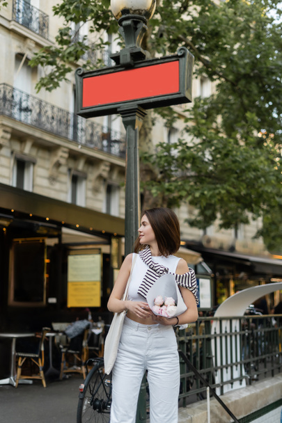 стильная женщина, улыбающаяся и держащая букет пионов, завернутых в бумагу, стоя на городской улице в Париже  - Фото, изображение