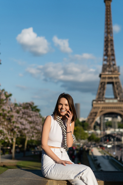 happy young woman in stylish outfit talking on smartphone while sitting near eiffel tower in paris, france - Zdjęcie, obraz