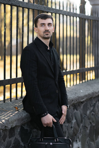 Een jongeman in een zwart pak staat bij het hek. Straatportret - Foto, afbeelding