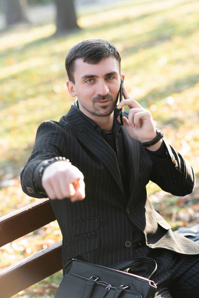 Młody brodaty mężczyzna rozmawia przez telefon i wskazuje palcem na kamerę. Biznesmen gestykulujący podczas rozmowy przez telefon - Zdjęcie, obraz
