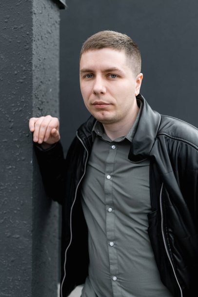 Un jeune homme vêtu d'une veste noire et d'une chemise verte regarde la caméra debout près du mur. Portrait de rue - Photo, image
