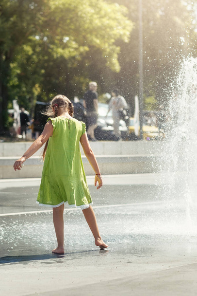 A young girl in a light green dress barefoot dances by a fountain in the city center. - Photo, Image