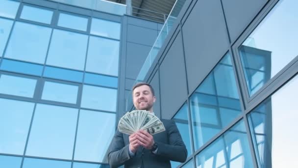 幸せな顔をしたクレイジービジネスマンはお金を散乱。人々はアメリカの通貨を使うでしょう。新しいビジネスセンターの背景に彼の手にドルを持つ男. - 映像、動画