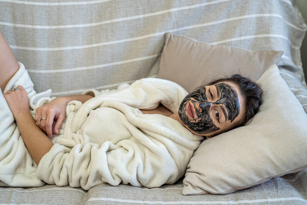 girl on the couch with a cosmetic mask made of clay on her face relaxes. The concept of a girl in a cosmetic mask - Foto, Bild