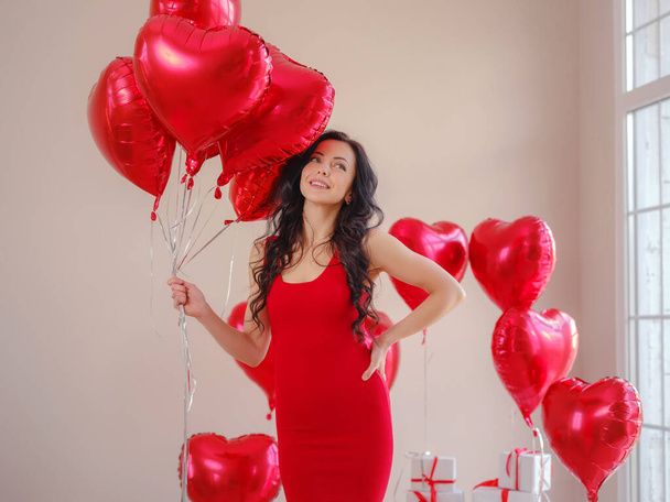 Όμορφη ευτυχισμένη νεαρή γυναίκα με κόκκινο φόρεμα. Πάρτι διακοπών. Χαρούμενο μοντέλο ποζάρει με μπαλόνια σε σχήμα κόκκινης καρδιάς, διασκεδάζει, γιορτάζει την ημέρα του Αγίου Βαλεντίνου. Σύμβολο αγάπης - Φωτογραφία, εικόνα