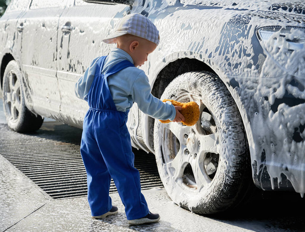 Kleiner Junge, der neben dem Auto steht, mit Schaum bedeckt, hält Schwamm in der Hand und lernt Autowaschen. Männliches Kind lernt Auto mit Schwamm waschen und beugt sich zum Vorderrad. - Foto, Bild